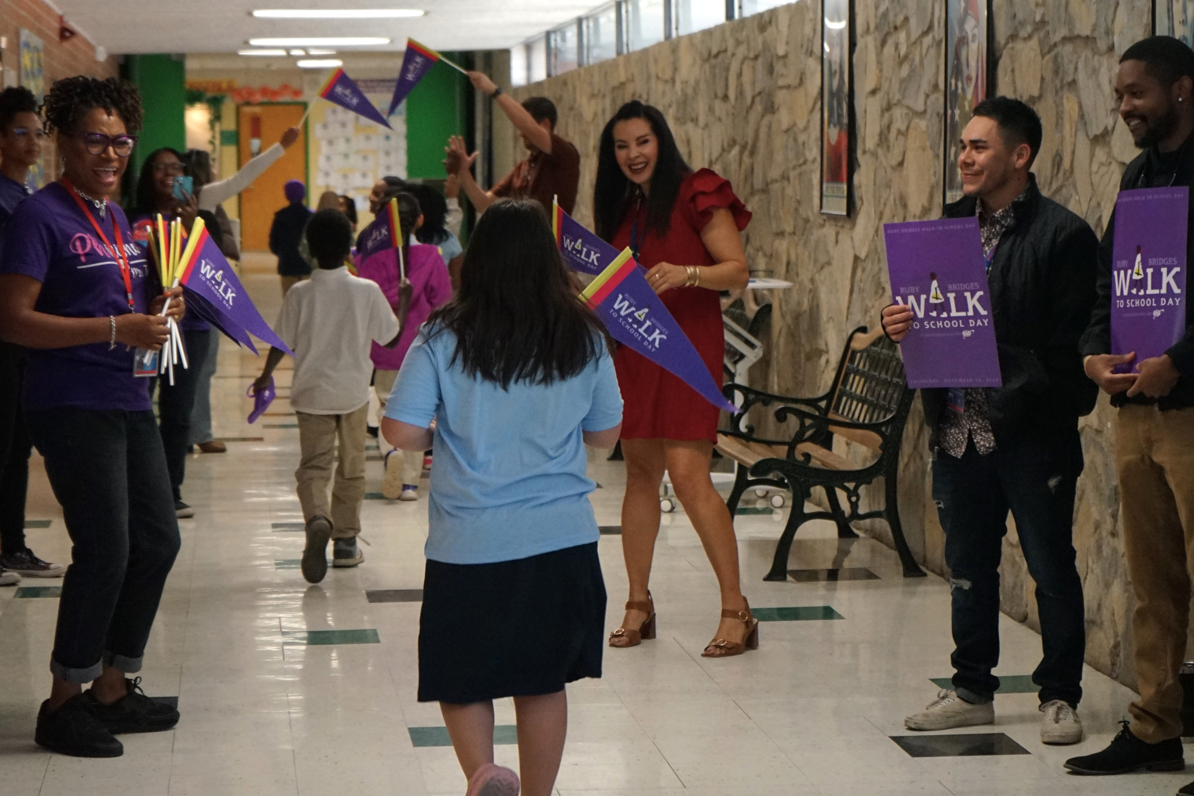 A girl in a blue shirt walks down the hallway with a purple flag as adults cheer her on