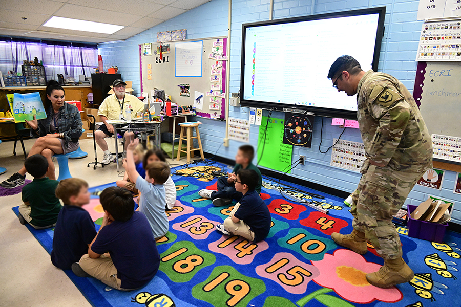 A man in military uniform sneaks up behind his son while he and his classmates are sitting on the floor listening to a book reading