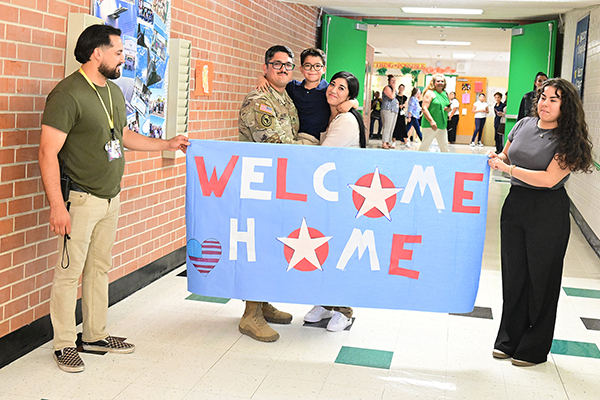 A man and woman hold up a blue banner with the words Welcome Home and a man in a military uniform, a woman and their young son stand behind the banner