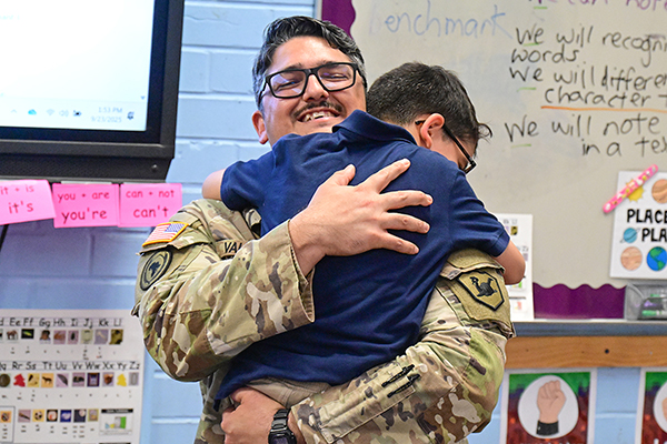 A man in a military uniform and glasses smiles as he hugs his young son