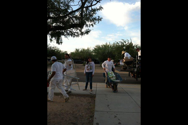 Men unload truck while another pushes wheelbarrow with supplies. Woman stands on sidewalk while men walk by with chairs