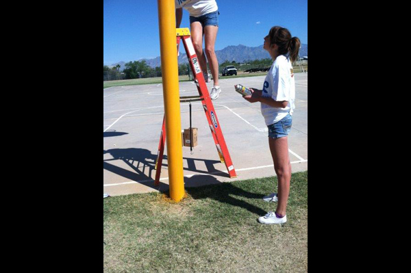 Woman holds pray paint while another climbs on to ladder beside basketball goal on a basketball court