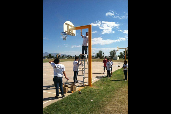 Man stands on ladder fixing basketball goal on basketball court while others watch
