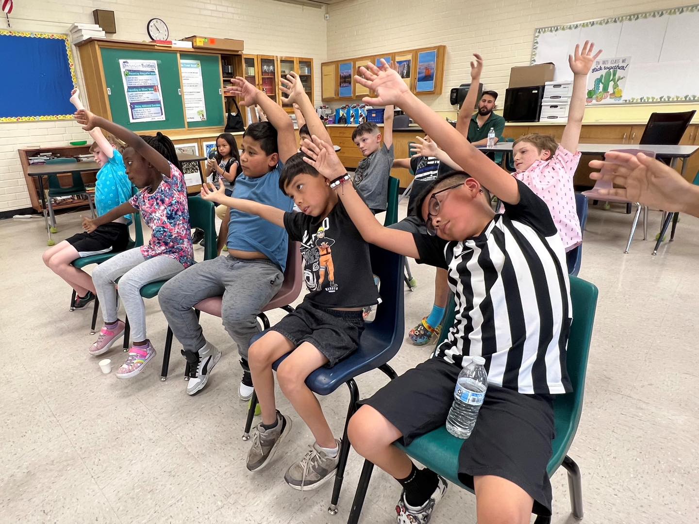 Students sitting in chairs wave their hands to the left during a summer school activity