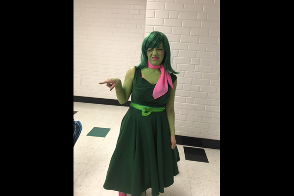 Student with green painted skin and wearing a green dress makes face and points in hallway