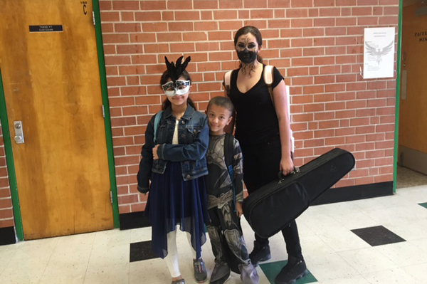 3 students stand in hall. One is wearing a mask with feathers, one is dressed in plain clothes, and the other is dressed in black with half her face painted black